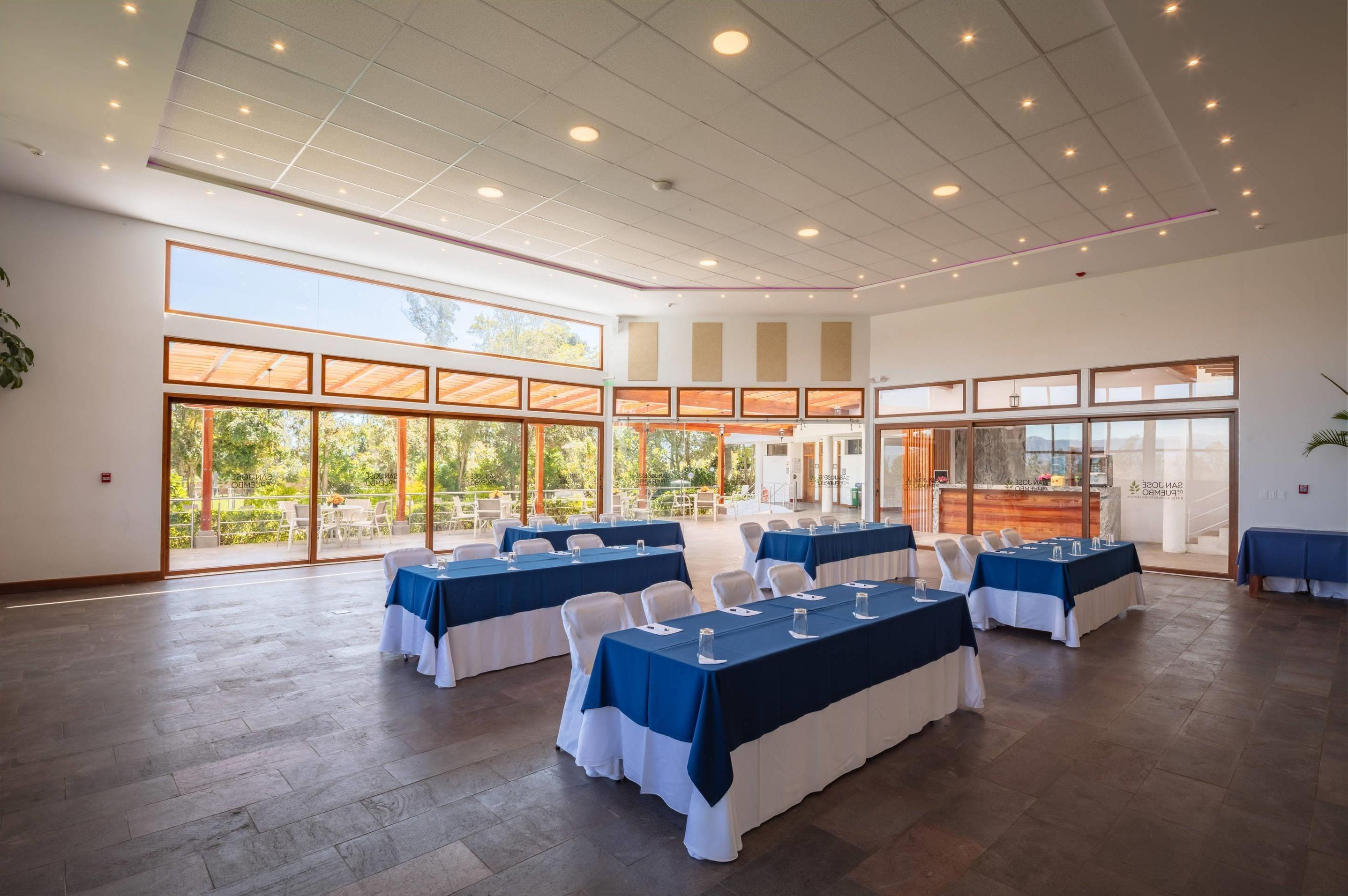 A spacious indoor area with multiple tables set up for dining, large windows offering views of the outdoors, and a modern ceiling design.