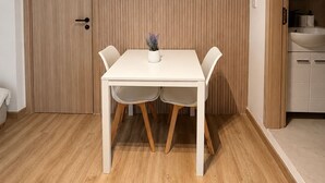Una habitación con piso de madera, una mesa blanca con dos sillas, una planta en maceta y una puerta de madera.