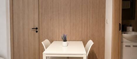 Ein Zimmer mit Holzboden, einem weißen Tisch mit zwei Stühlen, einer Topfpflanze und einer Holztür.