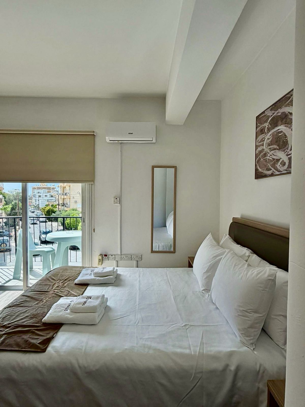 Hotelkamer met een bed, een raam met uitzicht op gebouwen, een airconditioning, een spiegel en een tafel met stoelen buiten.