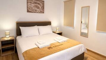 A neatly arranged hotel room with a bed, bedside tables, an air conditioning unit, a wall-mounted mirror, and a piece of wall art.