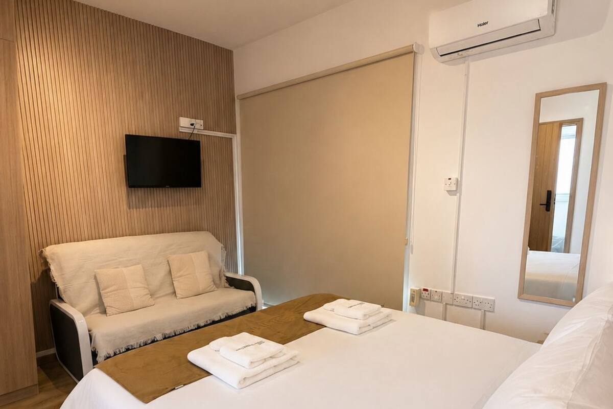 A hotel room with a large bed, a sofa, a flat-screen TV, and an air conditioning unit.