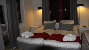 Cama doble con una manta roja, dos mesitas de noche, una lámpara y una ventana con cortinas.