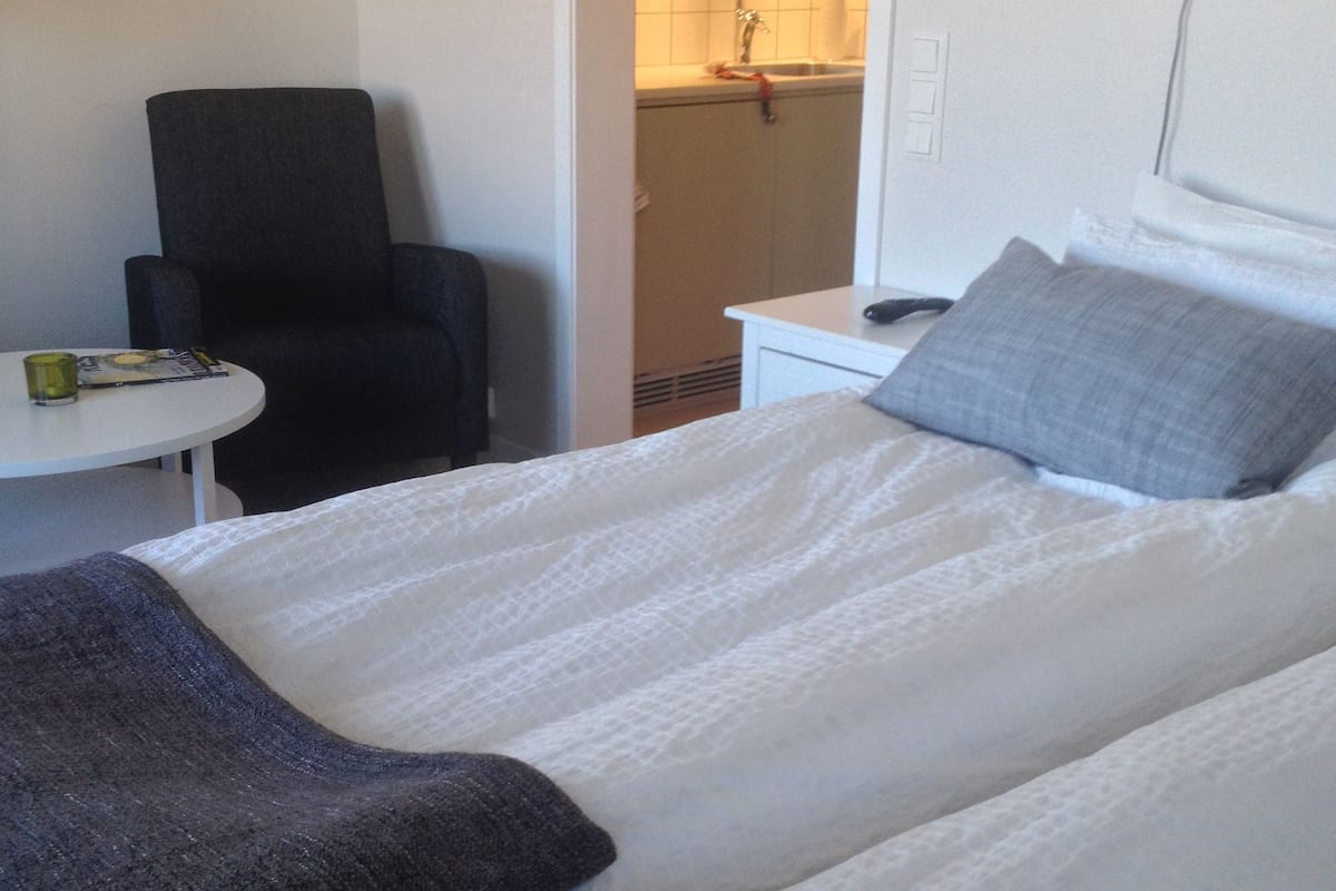 A hotel room with a bed, a chair, a small table, and a kitchenette in the background.