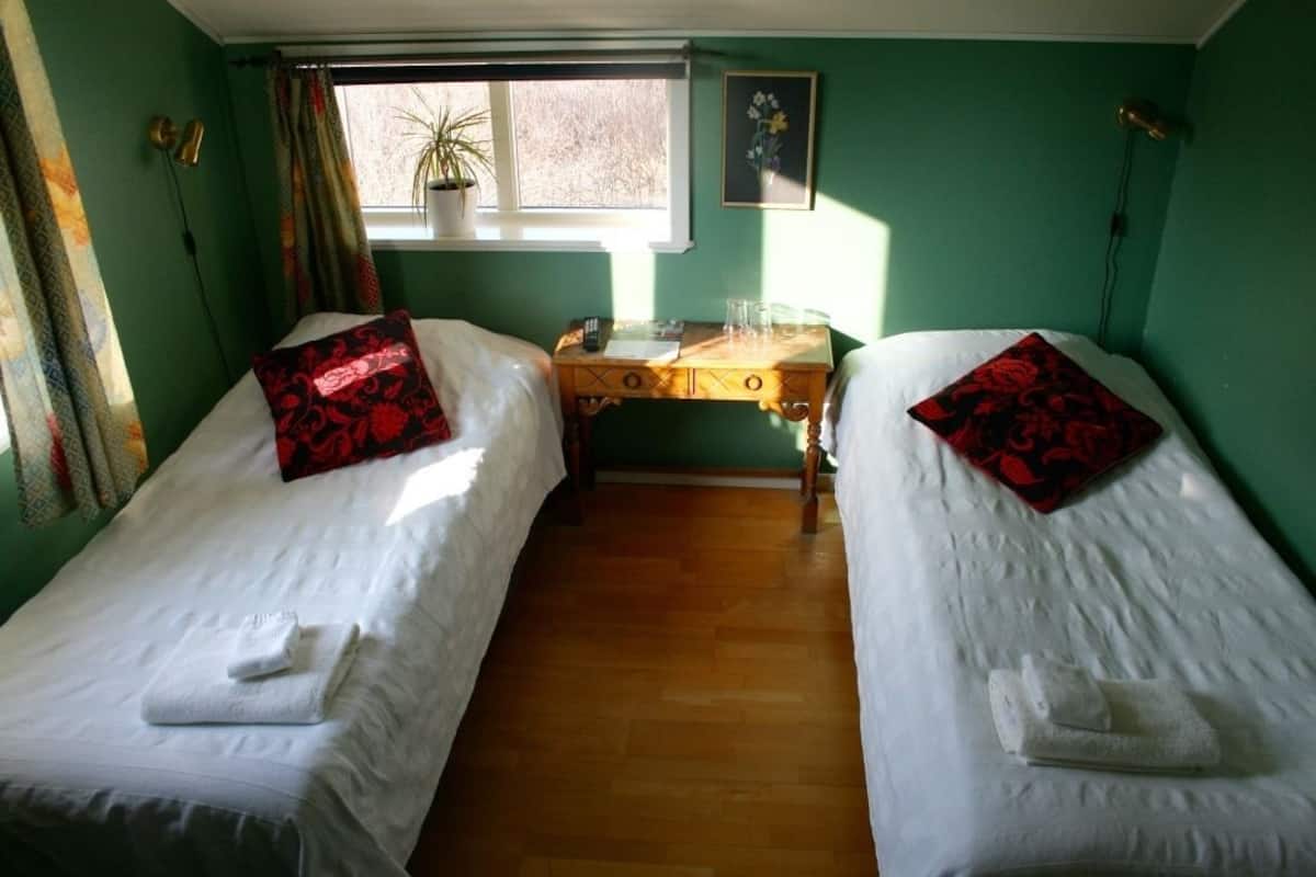 A room with two single beds, a wooden desk, a window with curtains, and a potted plant.