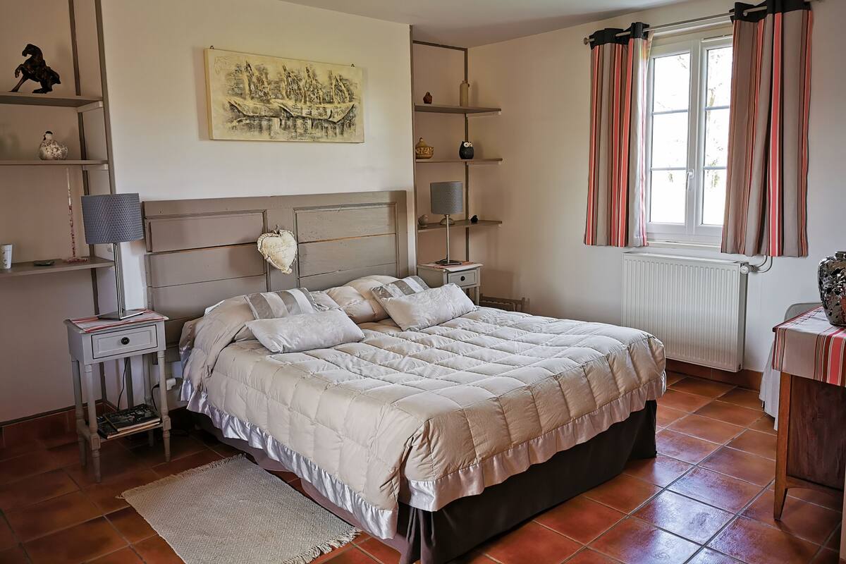 A bedroom with a bed, bedside tables, a nightstand, a lamp, a painting, a window with curtains, and a radiator.