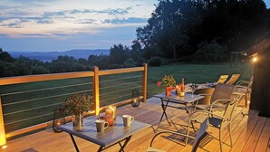 Terraza exterior con mesa lista para dos, sillas y barandilla con vista a un área con césped.