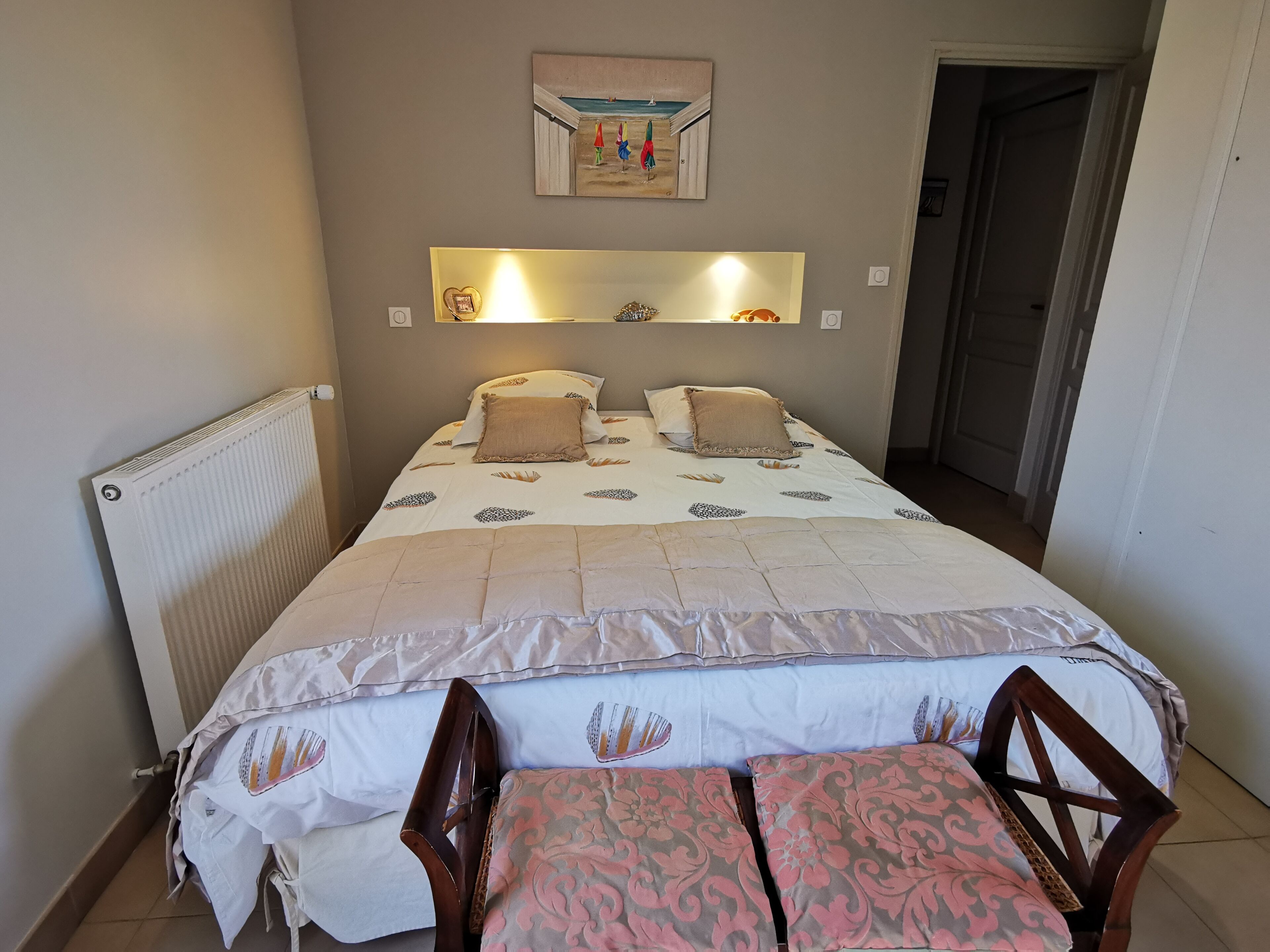 Una cama bien hecha con una colcha estampada y almohadas, una mesita de noche con una silla, un radiador y una estantería de pared con un cuadro.