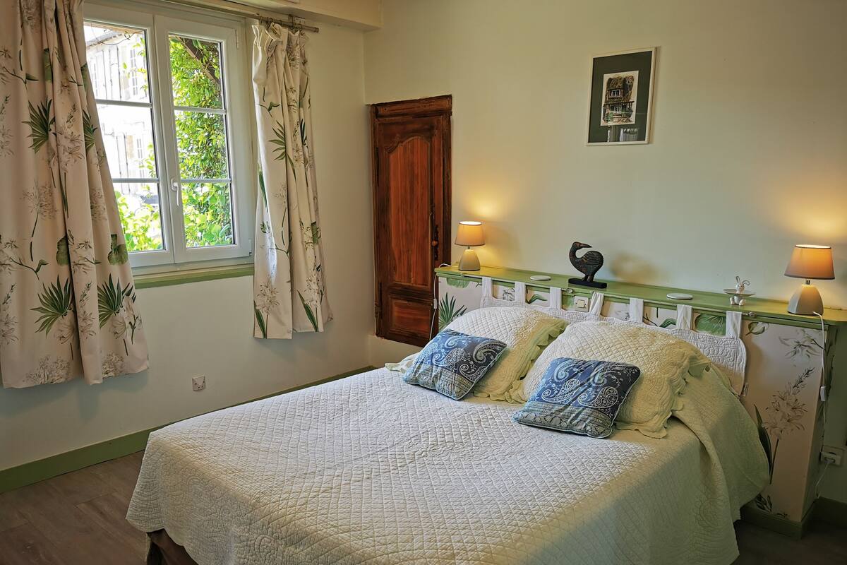 A bed with a quilted cover, two bedside lamps, a wooden headboard with a green top, and a window with curtains.
