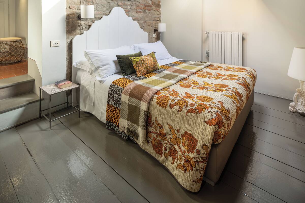 A bedroom with a stone wall, a wooden ceiling, a bed with a floral bedspread, a nightstand, and a lamp.
