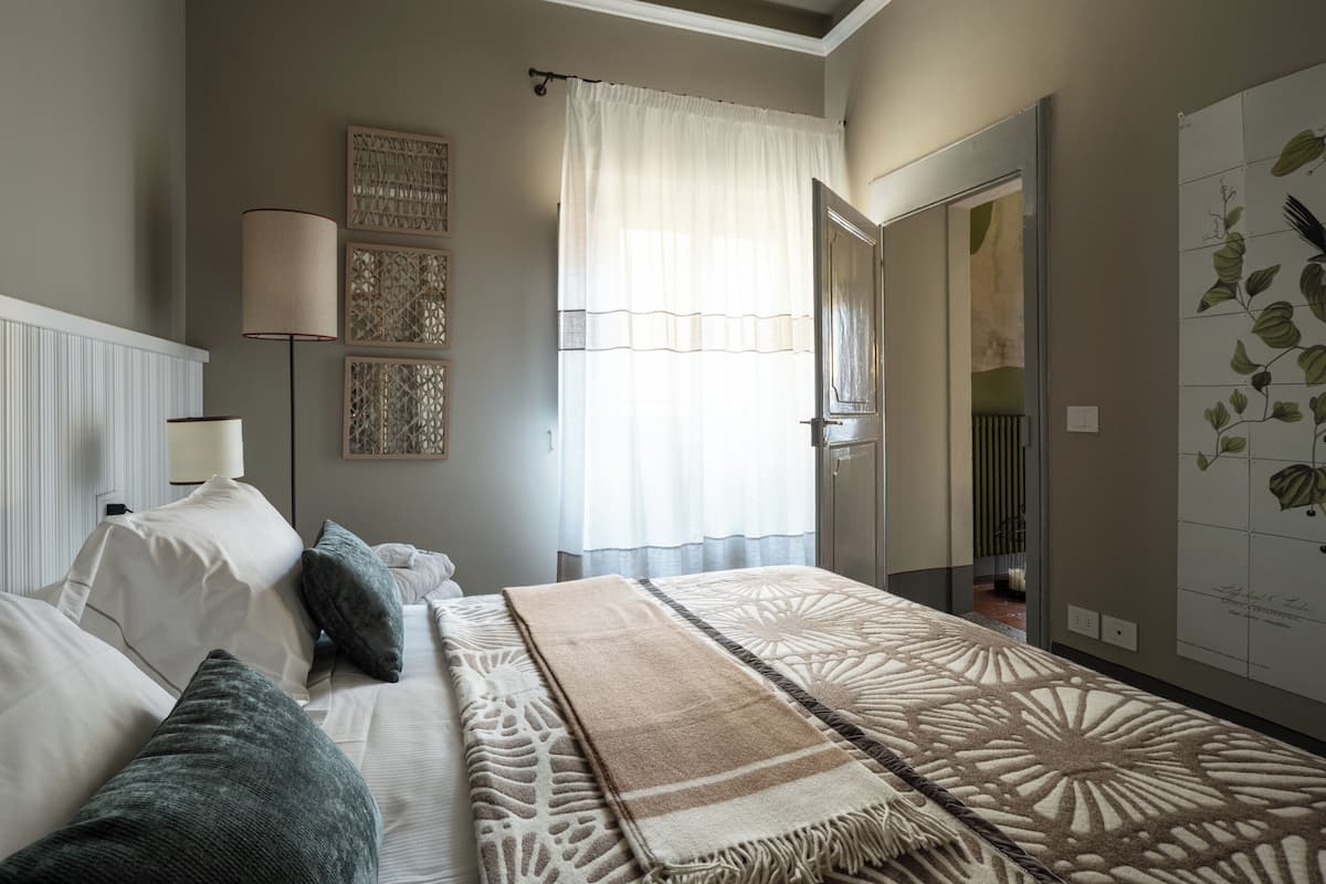 A bedroom with a bed, pillows, a blanket, a wall-mounted lamp, and a door leading to another room.