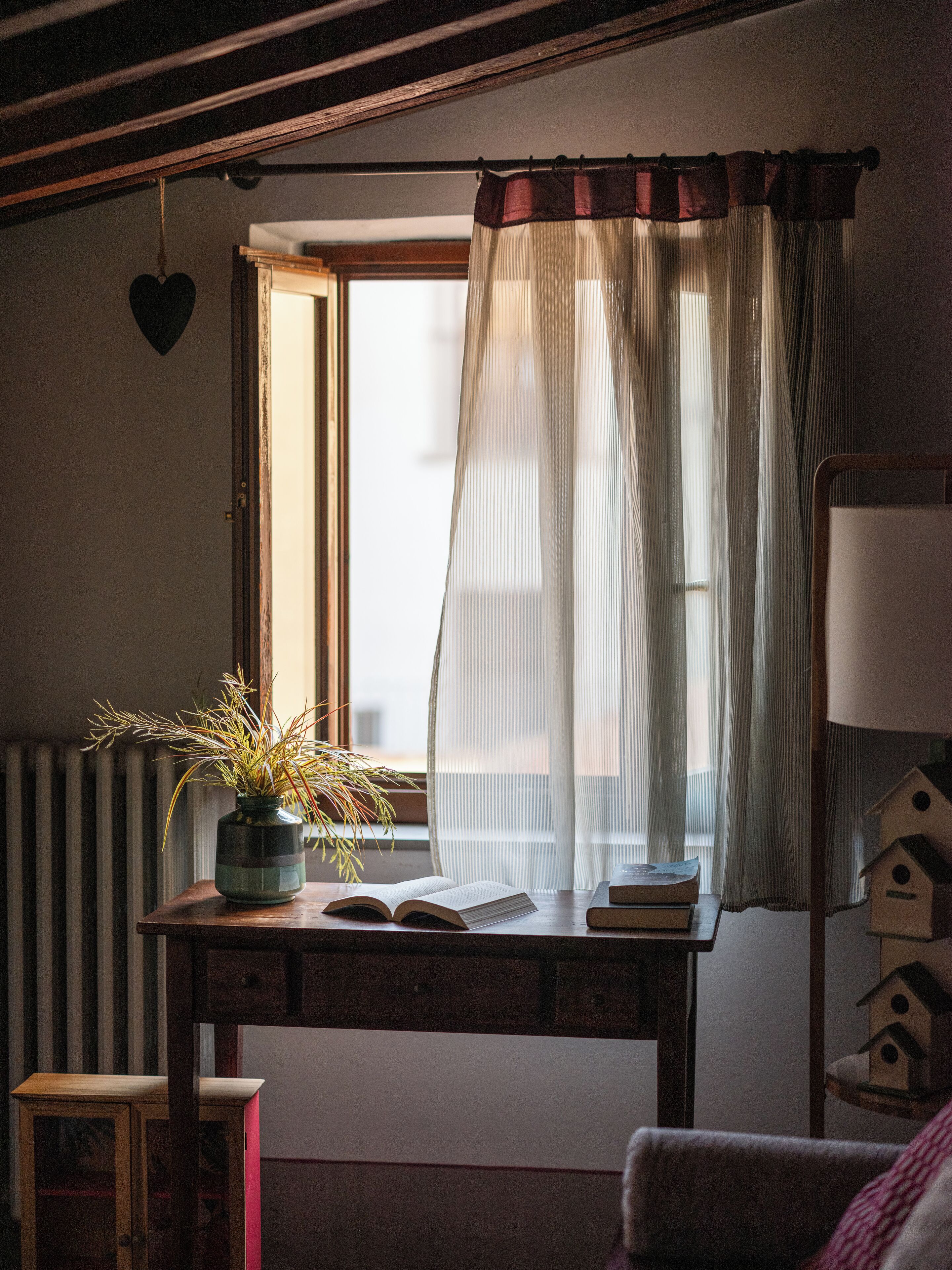A cozy room with a wooden desk, a potted plant, an open window with sheer curtains, a radiator, and a birdhouse.
