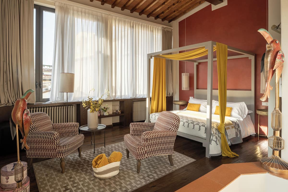 A hotel room with a four-poster bed, two patterned armchairs, a small table, a floor lamp, and a large window with curtains.