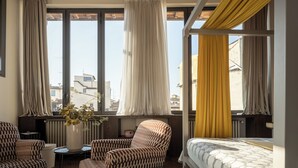 A modern hotel room with a patterned armchair, a sofa, a bed with a patterned duvet, a small table with a vase of flowers, and large windows with curtains. - Canto degli Scali (Florence)