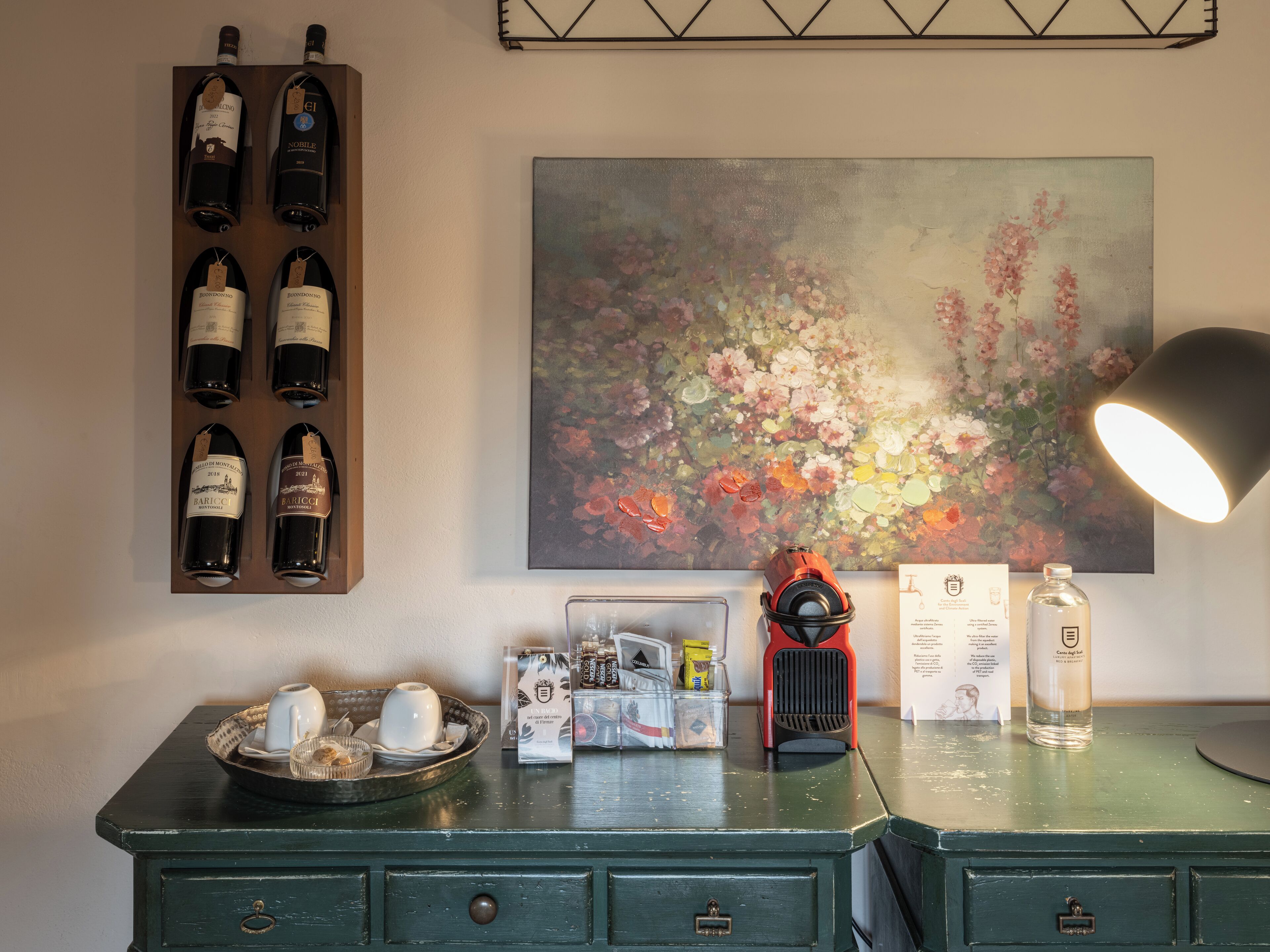 A room with a wine rack, a painting, a lamp, and a desk with various items.