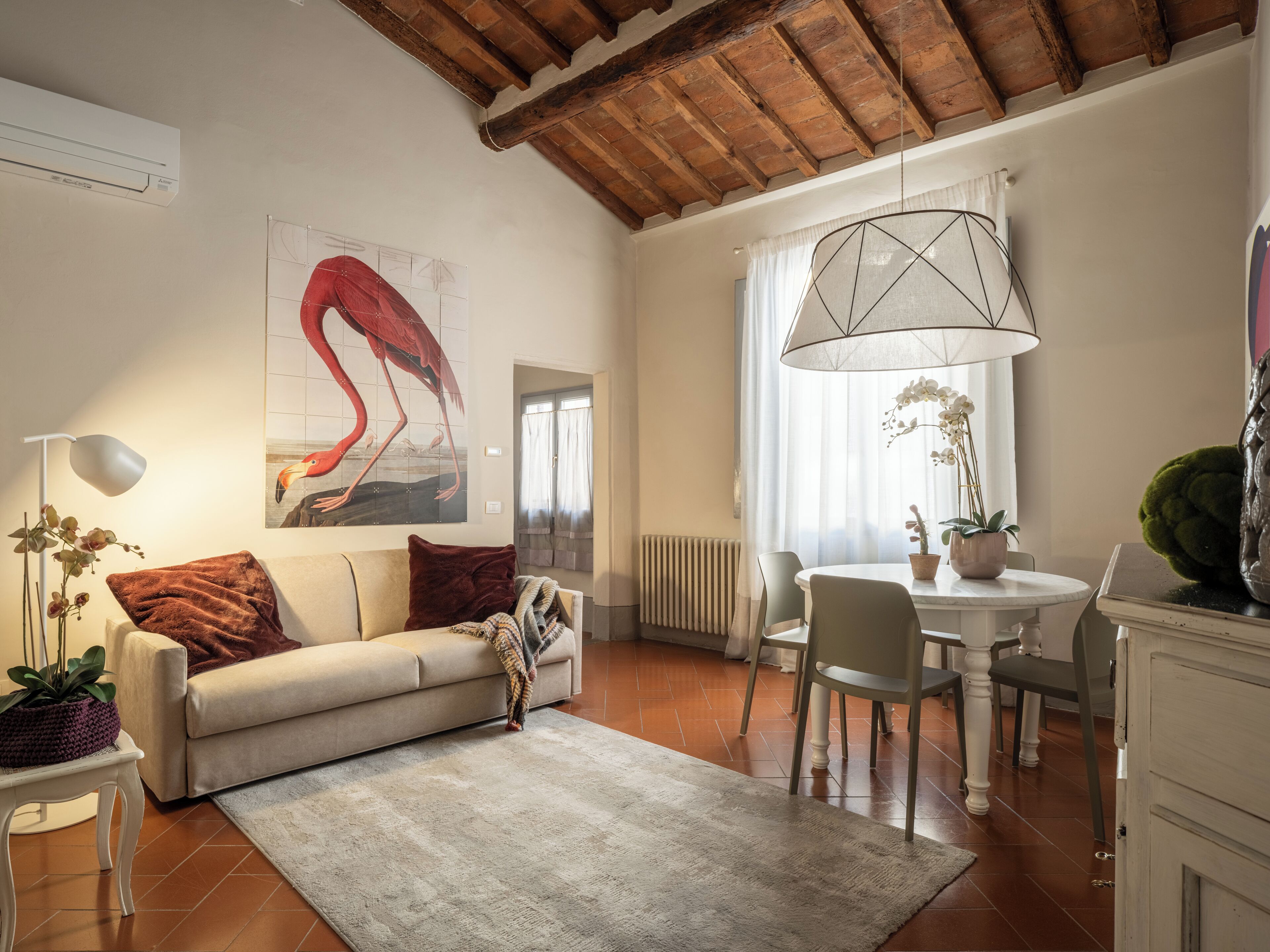 A living room with a sofa, a coffee table, a dining table with chairs, a hanging light fixture, and a radiator.