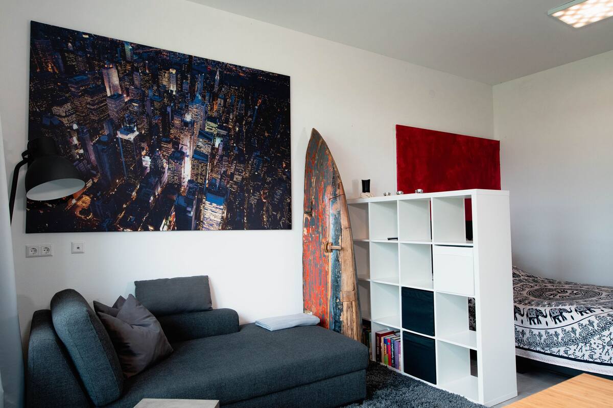 Un salon moderne avec un grand tableau représentant un paysage urbain, une planche de surf, un canapé, une bibliothèque et un lit à motifs.