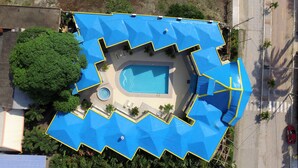 Eine Vogelperspektive eines Schwimmbadkomplexes mit einem markanten, blauen Dach, umgeben von viel Grün und nahe einer Straße.