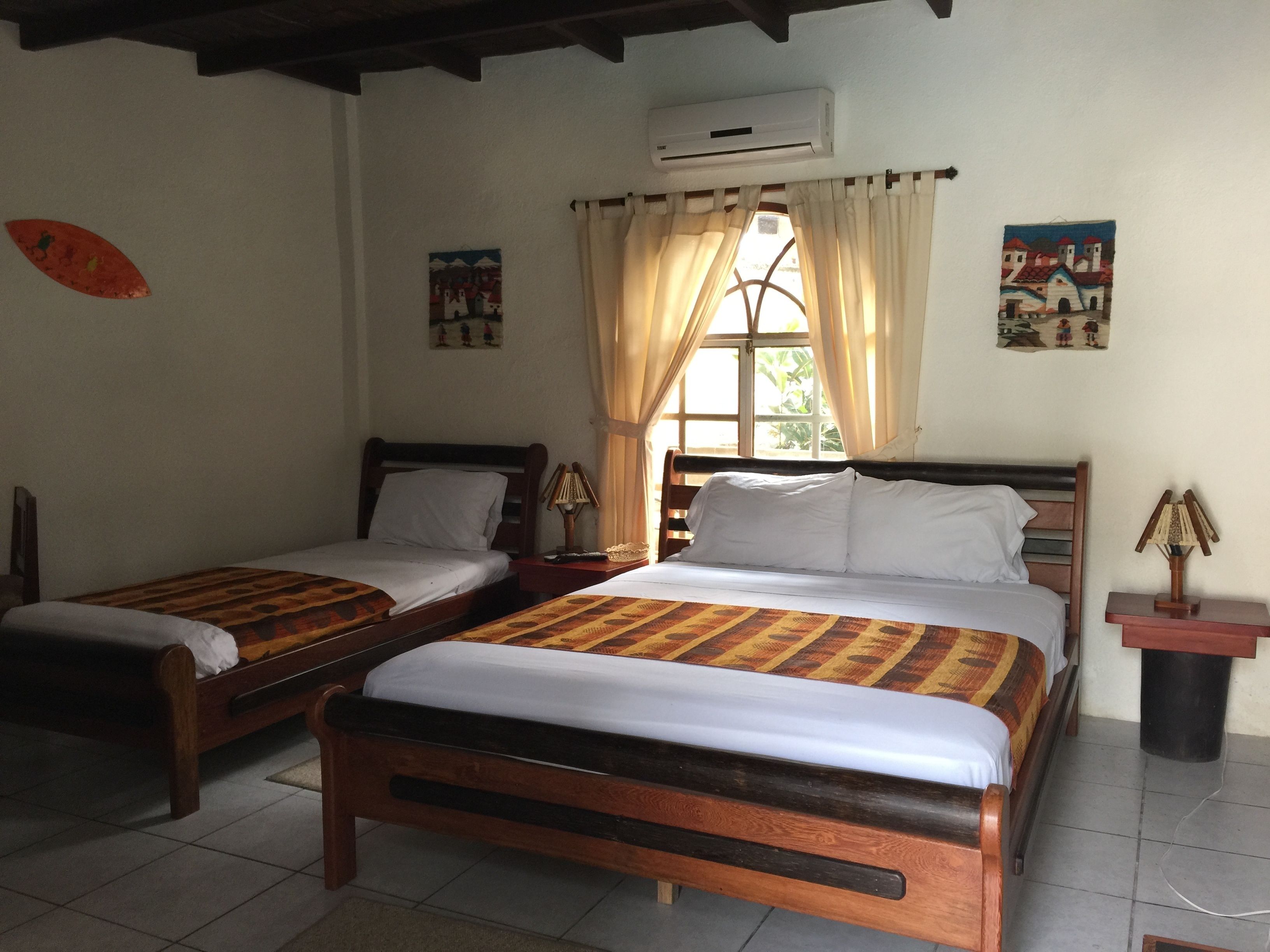 Una habitación con dos camas de madera, una ventana con cortinas, un equipo de aire acondicionado y una mesita con una lámpara.
