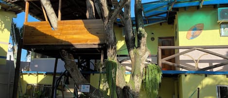 Une structure en forme de cabane dans les arbres, adossée à un bâtiment, et accessible par un escalier.