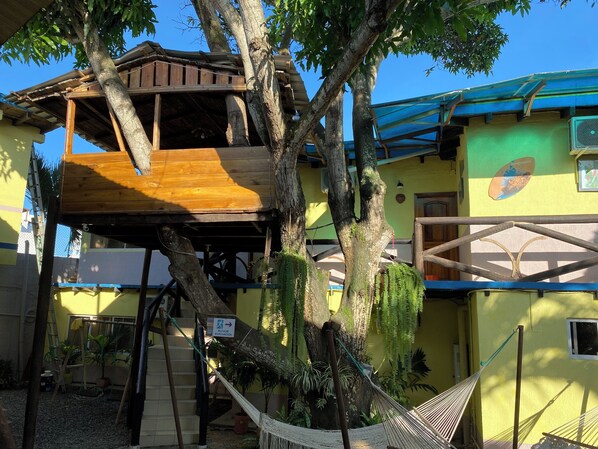 Une structure en forme de cabane dans les arbres, adossée à un bâtiment, et accessible par un escalier.