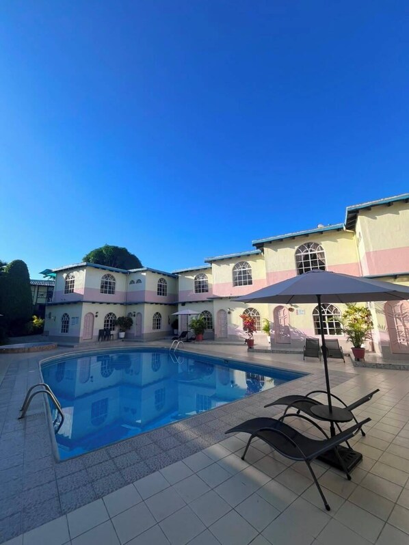 A swimming pool with lounge chairs and umbrellas, surrounded by a building with arched windows and a tiled floor.