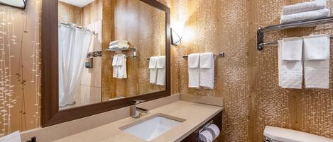 Un baño con lavamanos doble, un espejo grande y un inodoro. Hay toallas colgadas en un perchero y se ve una cortina de ducha en el espejo.