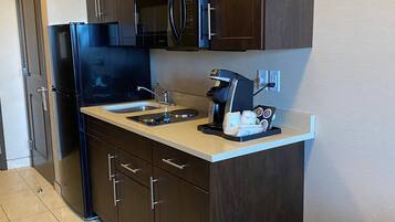 A compact kitchen with dark wooden cabinets, a black refrigerator, a microwave, and a coffee maker. There are two trash bins, one blue and one gray, on the floor.