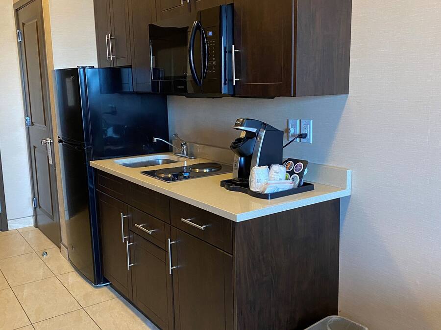 A compact kitchen with dark wooden cabinets, a black refrigerator, a microwave, and a coffee maker. There are two trash bins, one blue and one gray, on the floor.