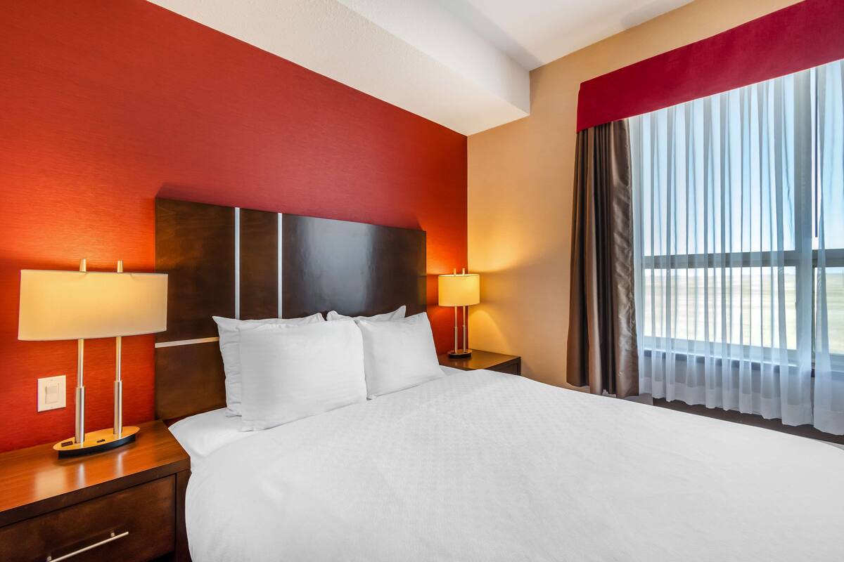 Een hotelkamer met een groot bed, twee nachtkastjes met lampen, een rode accentmuur en een raam met dunne gordijnen.