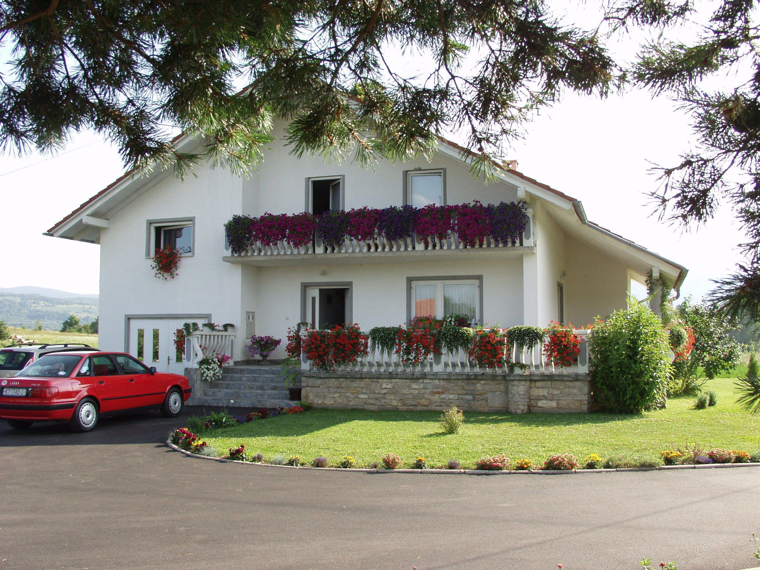 A two-story house with a white facade, a balcony with potted plants, and a red car parked in front.