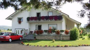 A two-story house with a white facade, a balcony with potted plants, and a red car parked in front.