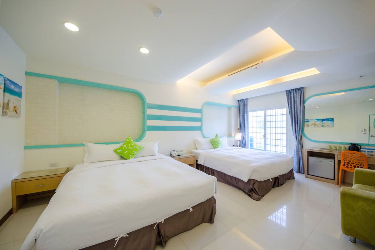 A hotel room with two beds, a desk, a chair, and a small kitchenette.
