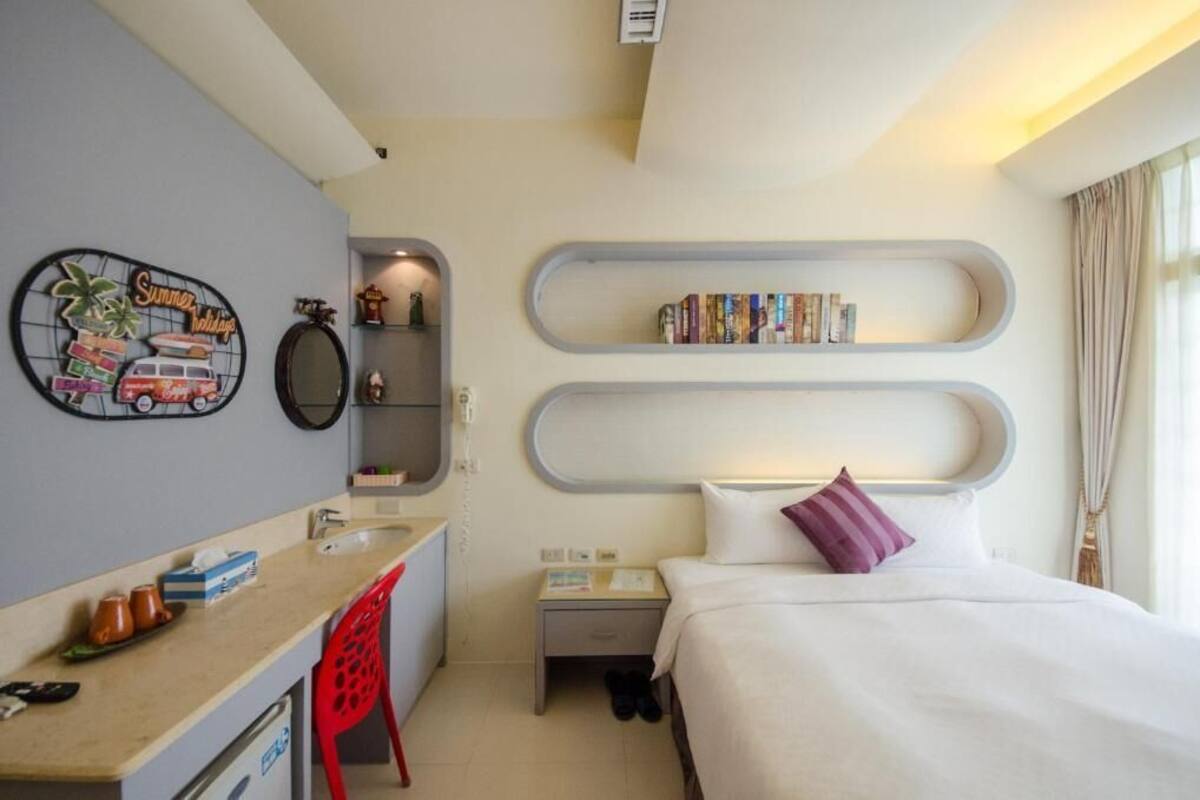 A compact hotel room with a bed, a small desk, a chair, a bookshelf, and a wall decoration.