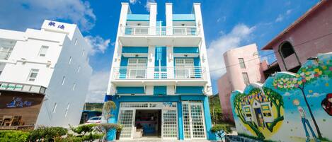 Un edificio de varios pisos con fachada azul y blanca, una entrada central y un mural en el lado derecho.