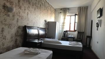 A hotel room with two beds, a wooden headboard, a radiator, a window with curtains, a small table with cups, and a chair.