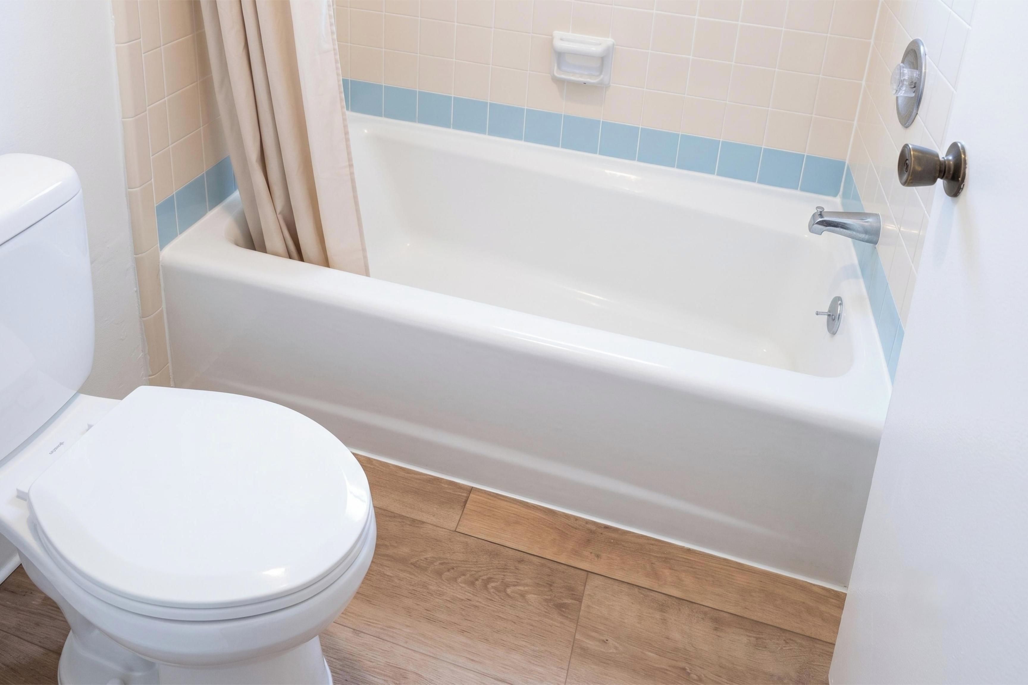 Un baño con inodoro blanco, bañera con detalle de azulejos azules y suelo de madera.