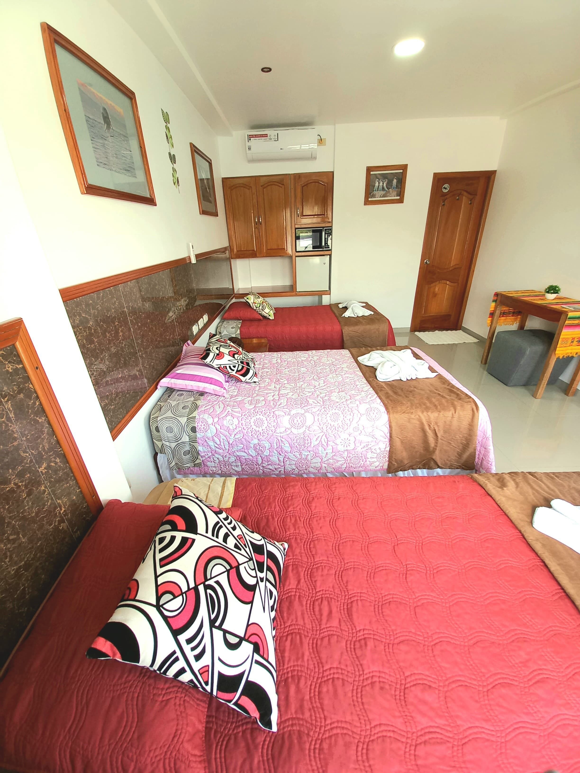 A hotel room with two beds, each with a red bedspread and patterned pillows. There is a wooden wardrobe, a small table with a plant, and an air conditioning unit on the wall.
