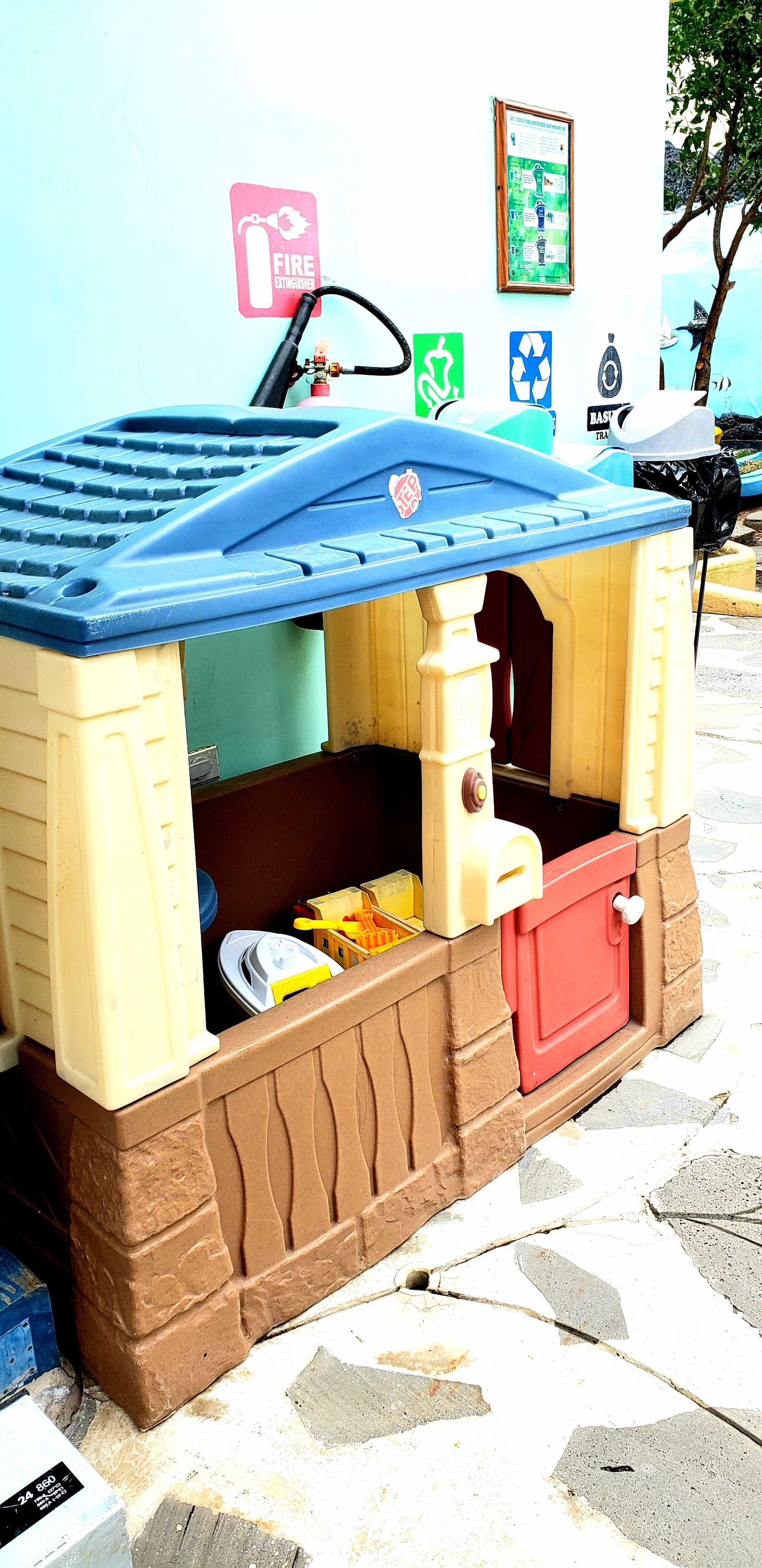 A children's playhouse with a blue roof and beige walls, situated on a tiled floor with a fire extinguisher sign in the background.