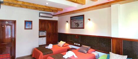 Habitación de hotel con vigas de madera en el techo, una cama con ropa de cama verde y marrón, un armario de madera y una pequeña mesa con un mantel colorido.