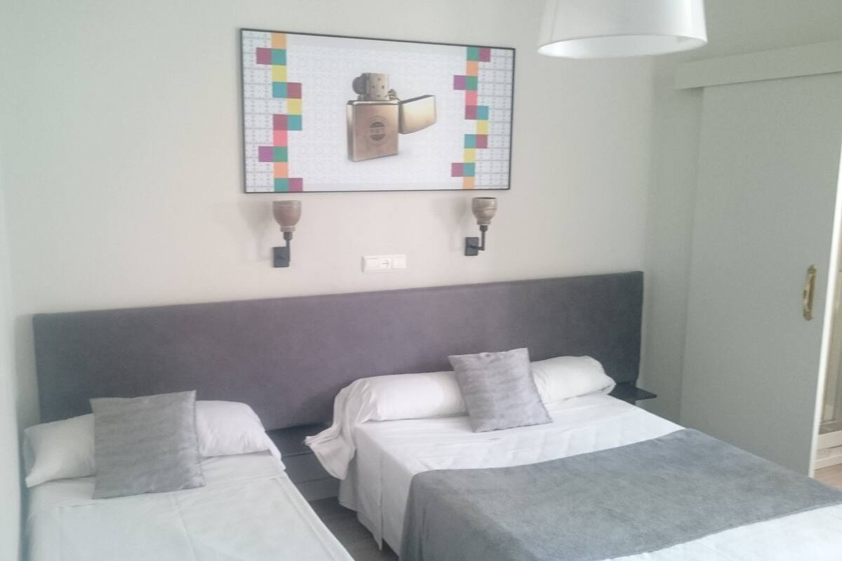 Habitación de hotel con dos camas individuales, cabecero gris y lámpara fijada a la pared.