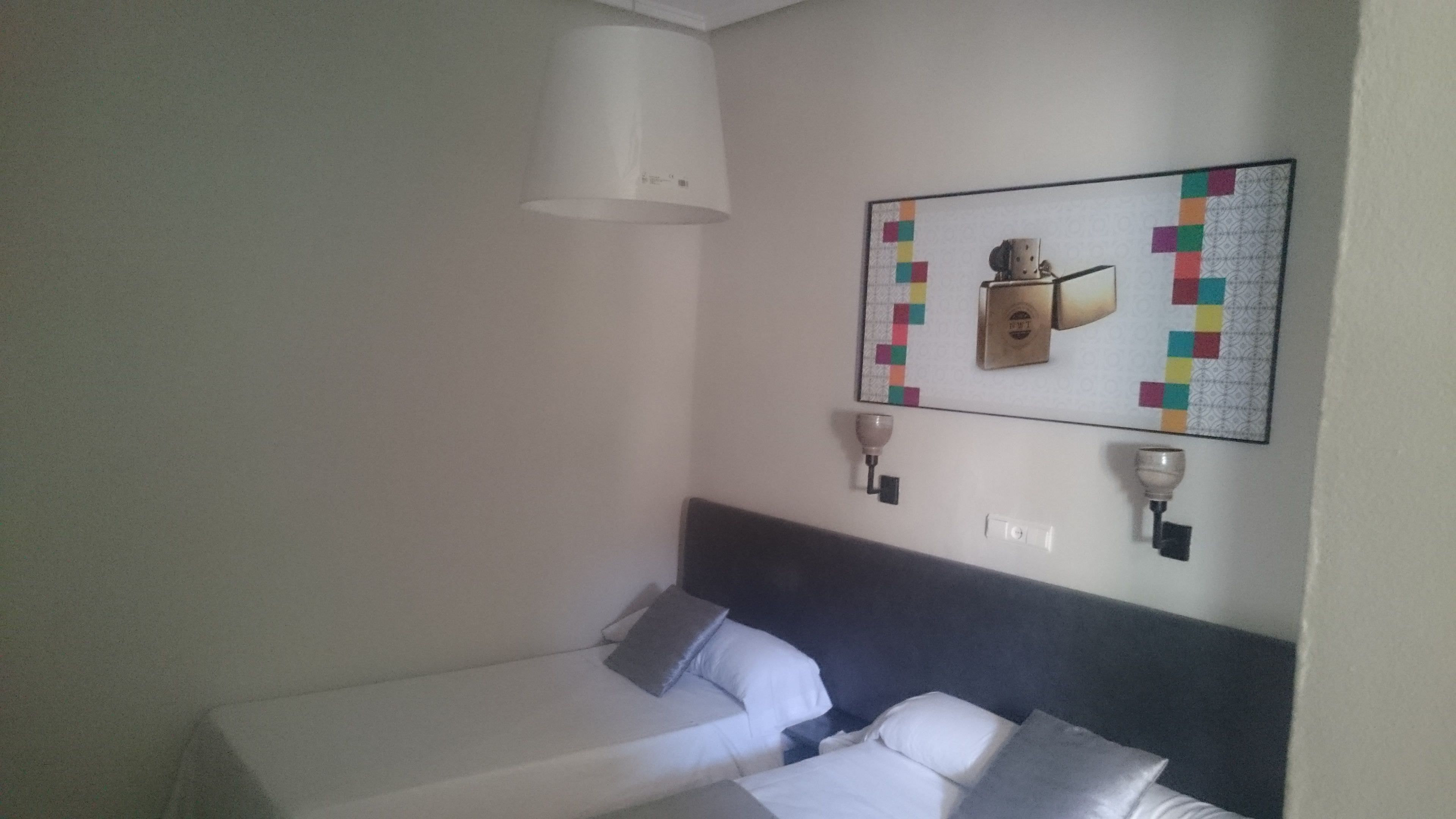 Habitación de hotel con dos camas individuales, cabecera, lámpara de pared y una obra de arte acolchada.