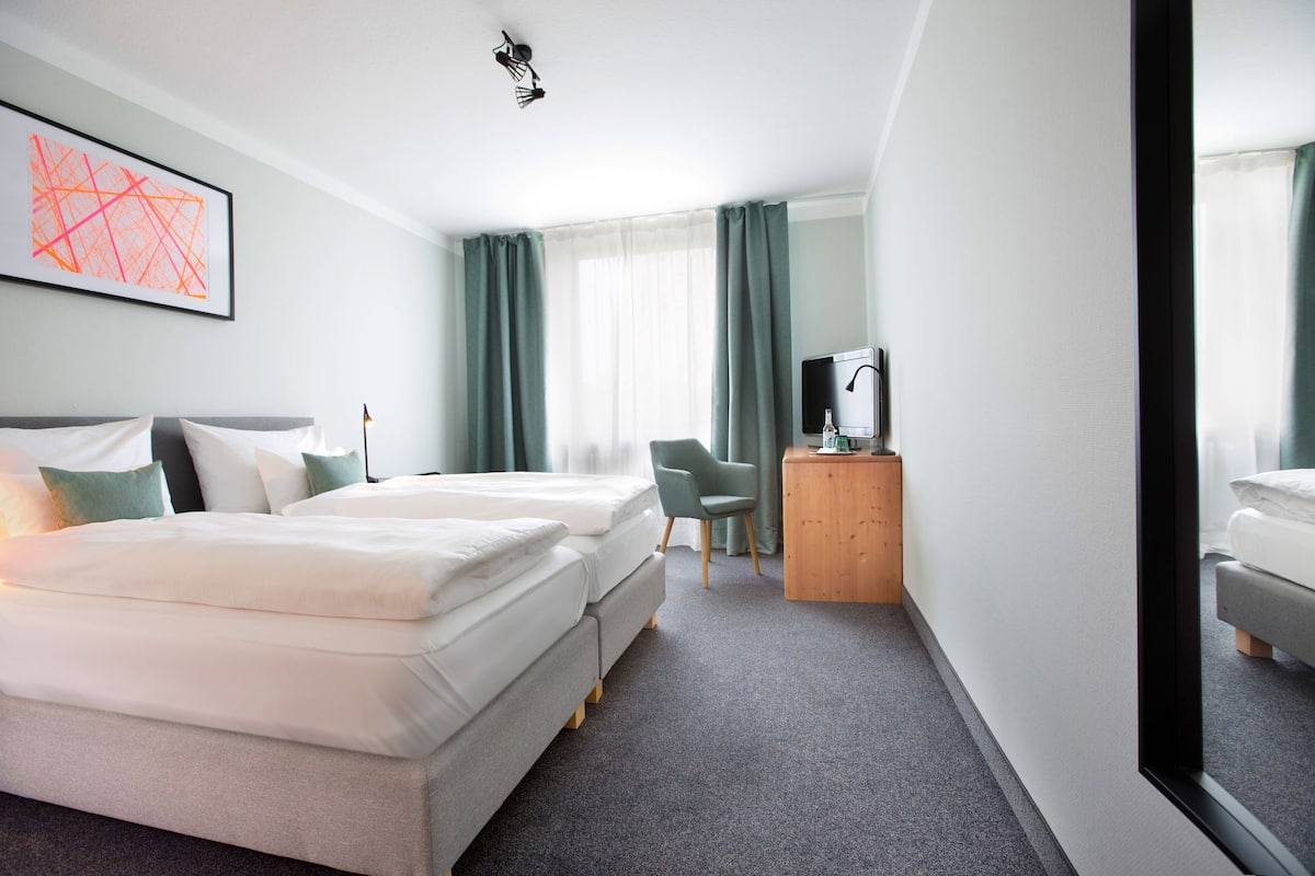 A hotel room with two beds, a desk with a chair, a TV, and a large window with curtains.