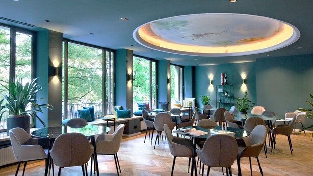 A modern dining area with round tables and chairs, large windows, and potted plants. - Hotel Rotkreuzplatz (Munich)