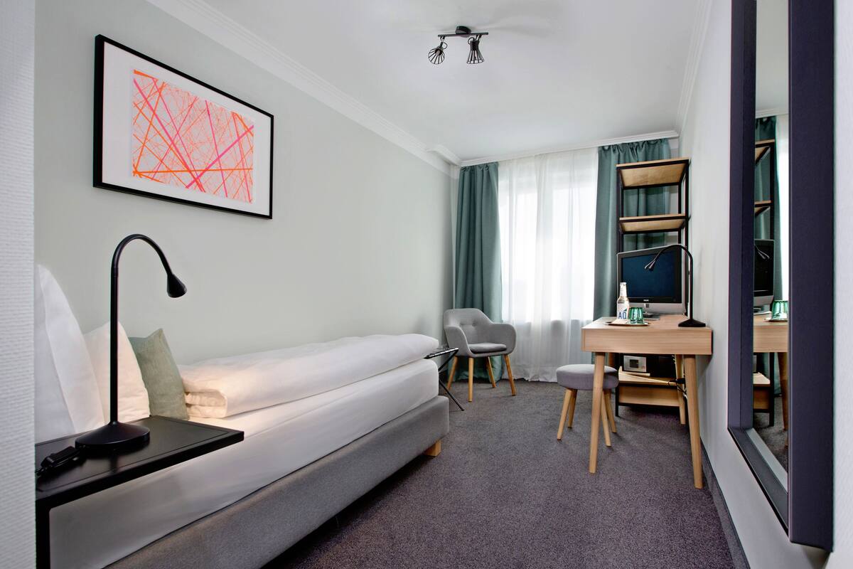 A modern hotel room with a single bed, a desk with a computer, a chair, and a bookshelf.