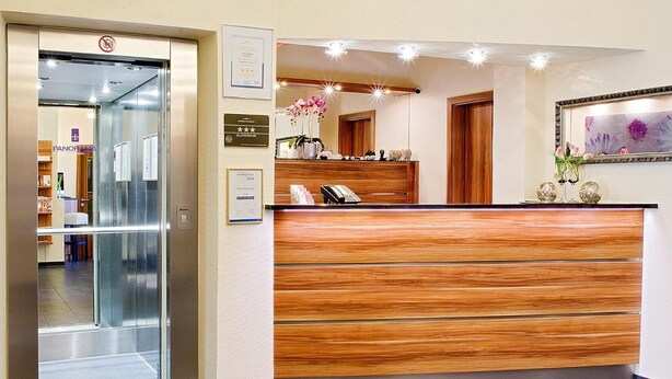A hotel reception area with a wooden reception desk, a glass door entrance, and a framed certificate on the wall.
