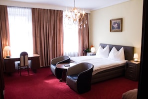 Une chambre d’hôtel avec un grand lit, un bureau, deux chaises, un lustre et un tableau au mur.