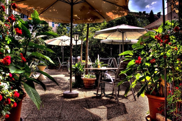 Une terrasse extérieure avec des tables et des chaises, entourée de plantes en pot et d’ombrelles.