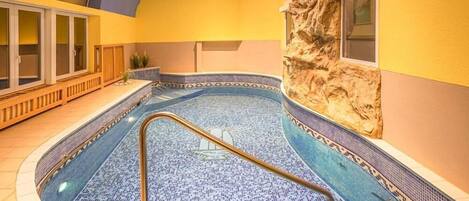 An indoor swimming pool with a tiled floor and a curved design, surrounded by wooden benches and a stone wall.