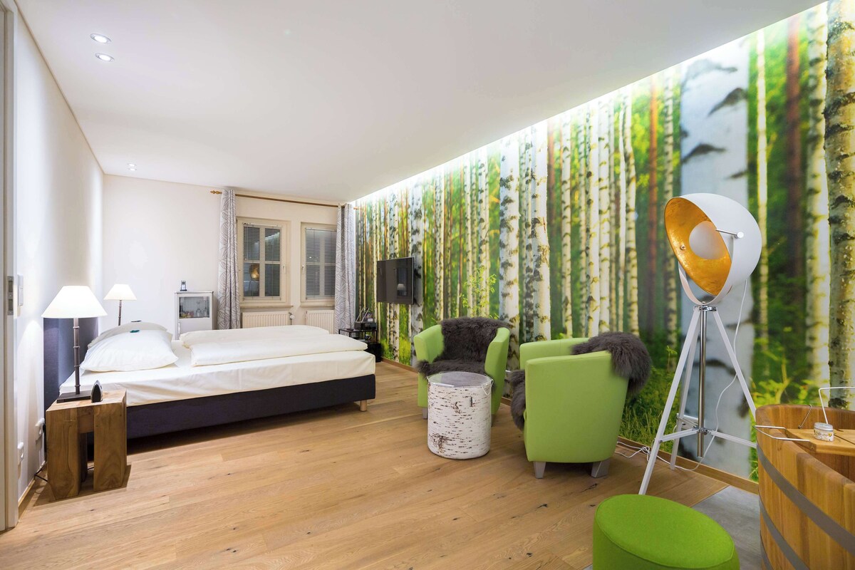 Un dormitorio moderno con cama, sillas verdes, una mesita de noche de madera y un mural en la pared.